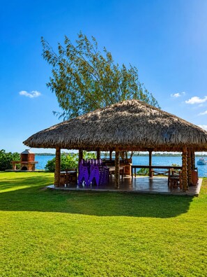 A thatched-roof pavilion with outdoor seating, a grill, and a view of a body of water.