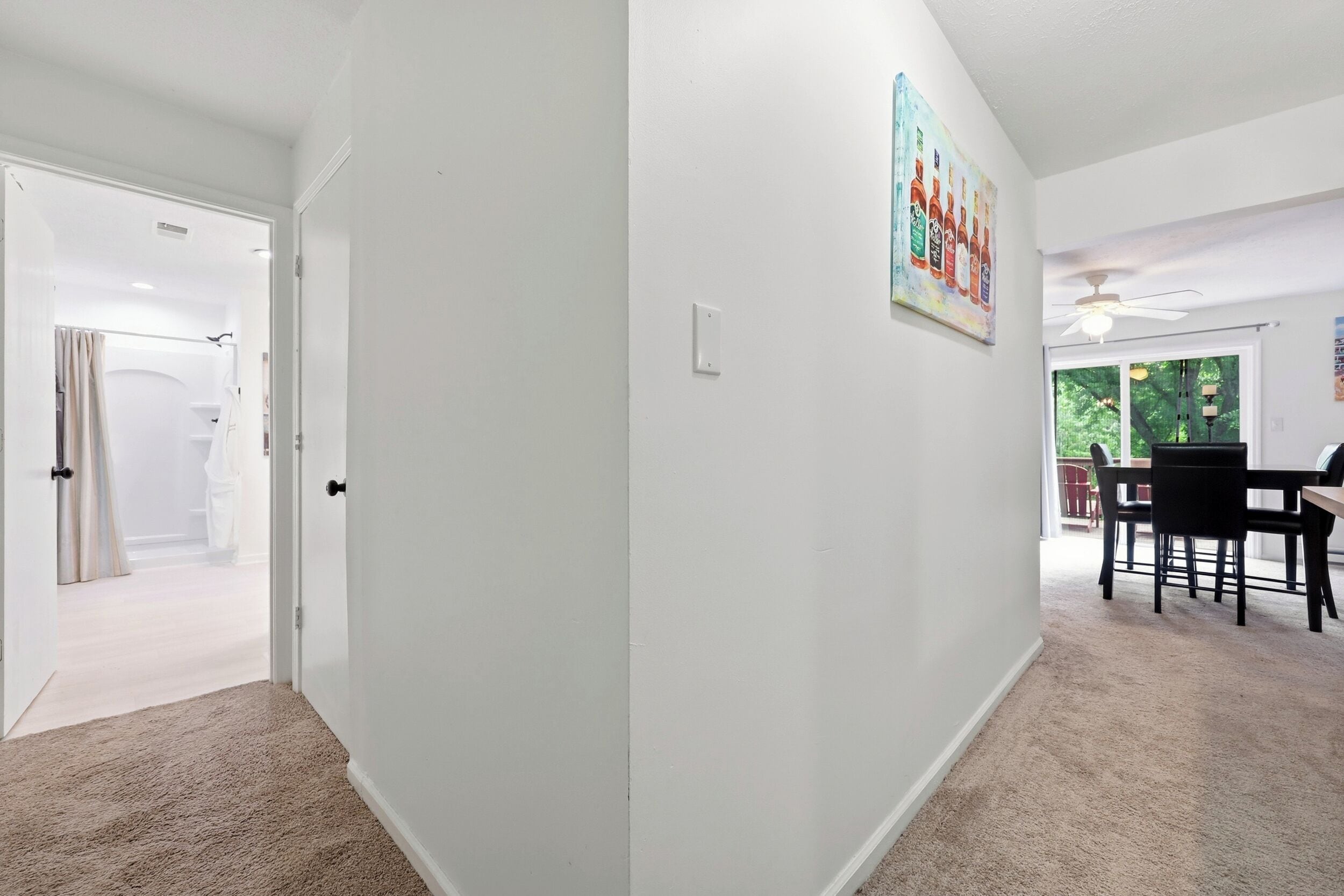 Um corredor com piso revestido de carpete, uma porta que dá para um banheiro e uma sala de jantar com mesa e cadeiras.