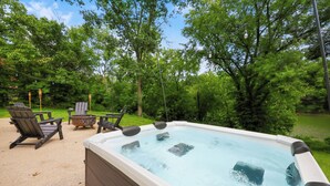 An outdoor hot tub surrounded by wooden deck chairs and a fire pit, set in a lush green garden. - River Bend Reserve (Frankfort)
