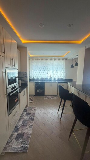 A modern kitchen with built-in appliances, a dining area with bar stools, and a patterned rug on the floor. - Élégance à Kouba – 3 chambres, suite & garage. (Kouba)