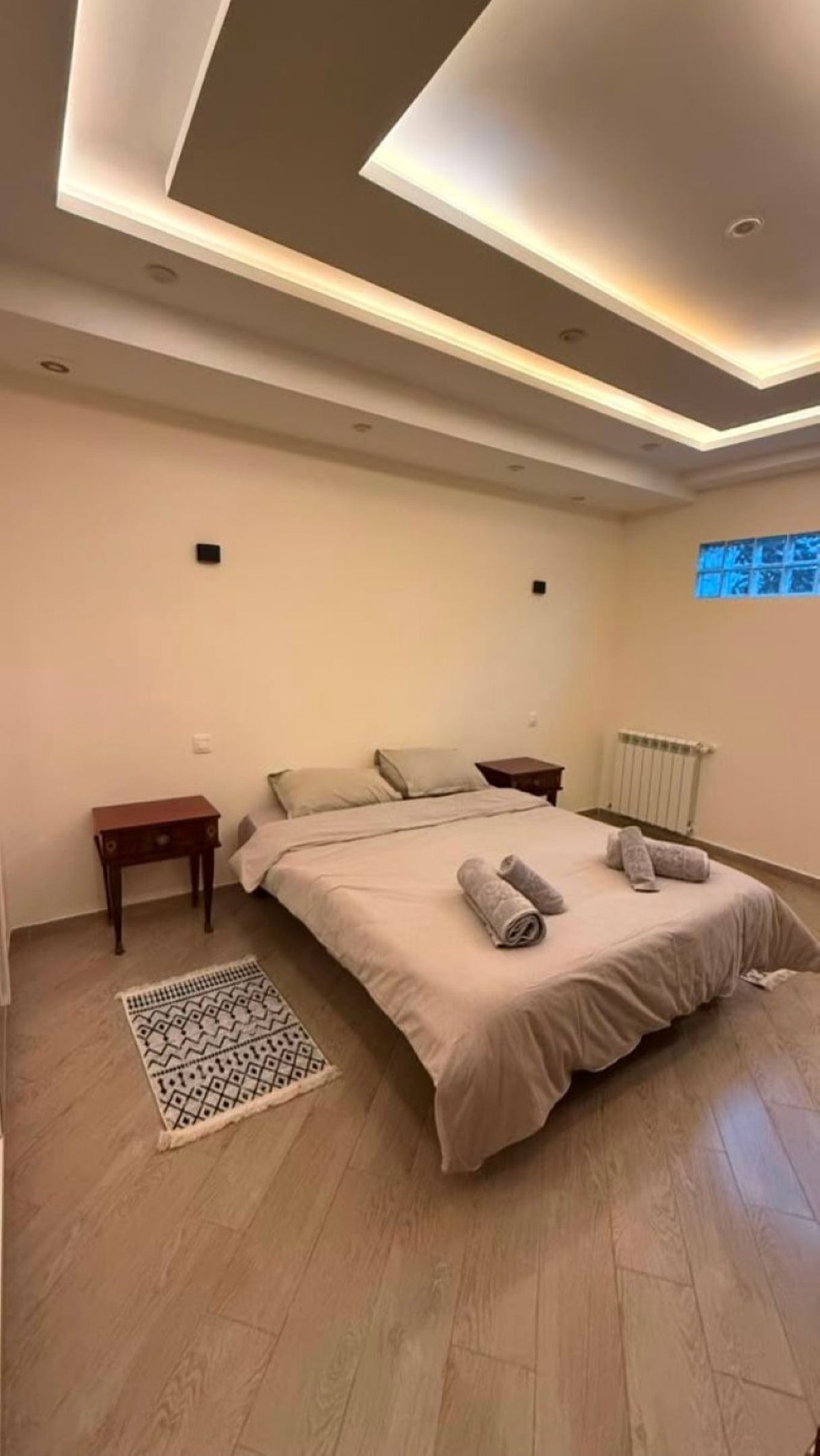 A modern bedroom with a large bed, bedside tables, a patterned rug, and a ceiling with recessed lighting.