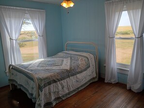 Una cama individual en una habitación con paredes azul claro y suelo de madera, con una colcha estampada y cortinas blancas.