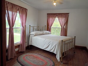 A bedroom with a single bed, checkered curtains, a patterned rug, and a view of greenery outside.