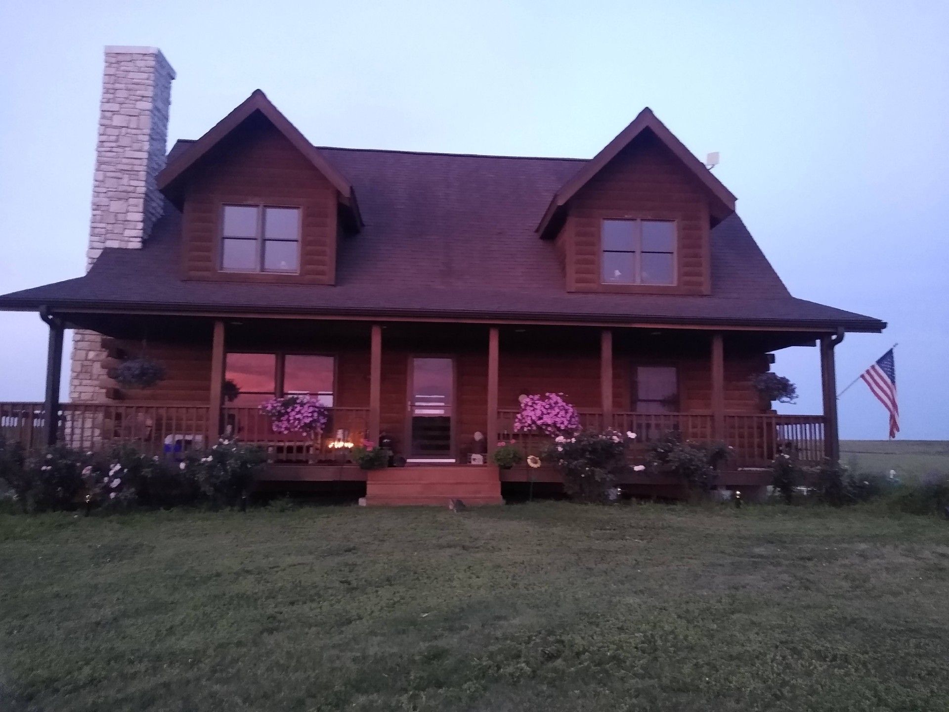 A two-story log cabin with a stone chimney, a front porch with flower arrangements, and an American flag.