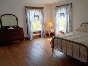 Un dormitorio con piso de madera, una cama con estructura de metal, un aparador con espejo y dos ventanas con cortinas a cuadros.