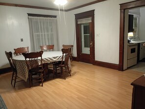 Comedor con mesa y sillas de madera, ventana con cortinas, y una cocina visible a través de una puerta abierta.