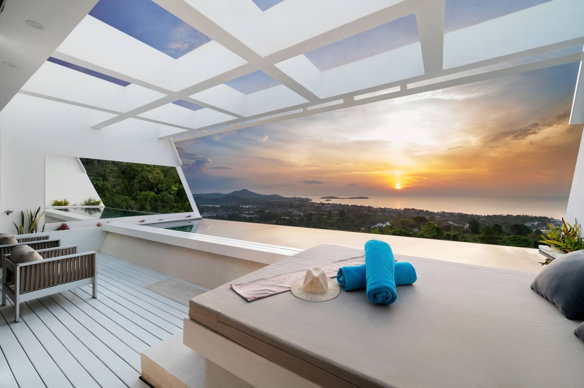 Une terrasse moderne sur le toit, équipée d’un ordinateur portable, d’un espace salon et offrant une vue sur le coucher de soleil au-dessus d’un paysage côtier.