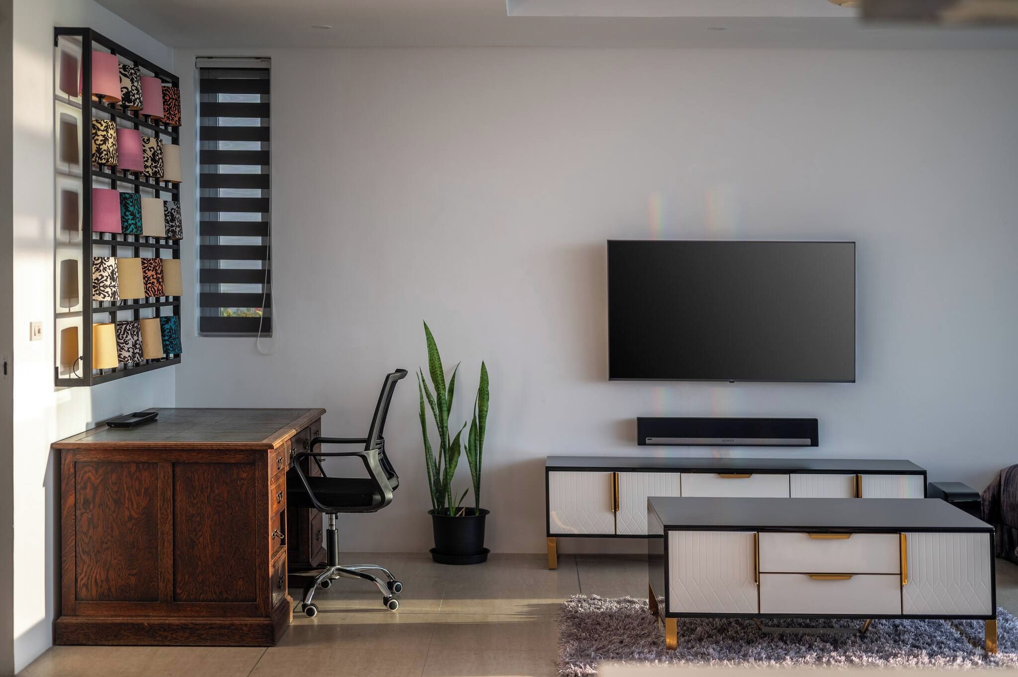 Uma sala de estar moderna com uma escrivaninha de madeira, uma cadeira de escritório preta, um vaso de planta, uma TV de tela plana e um aparador branco.
