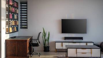 Uma sala de estar moderna com uma escrivaninha de madeira, uma cadeira de escritório preta, um vaso de planta, uma TV de tela plana e um aparador branco.