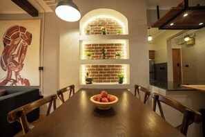 Ein Essbereich mit einem Holztisch, Stühlen und einer Obstschale. Im Hintergrund sind dekorative Lampen und eine Ziegelwand zu sehen.