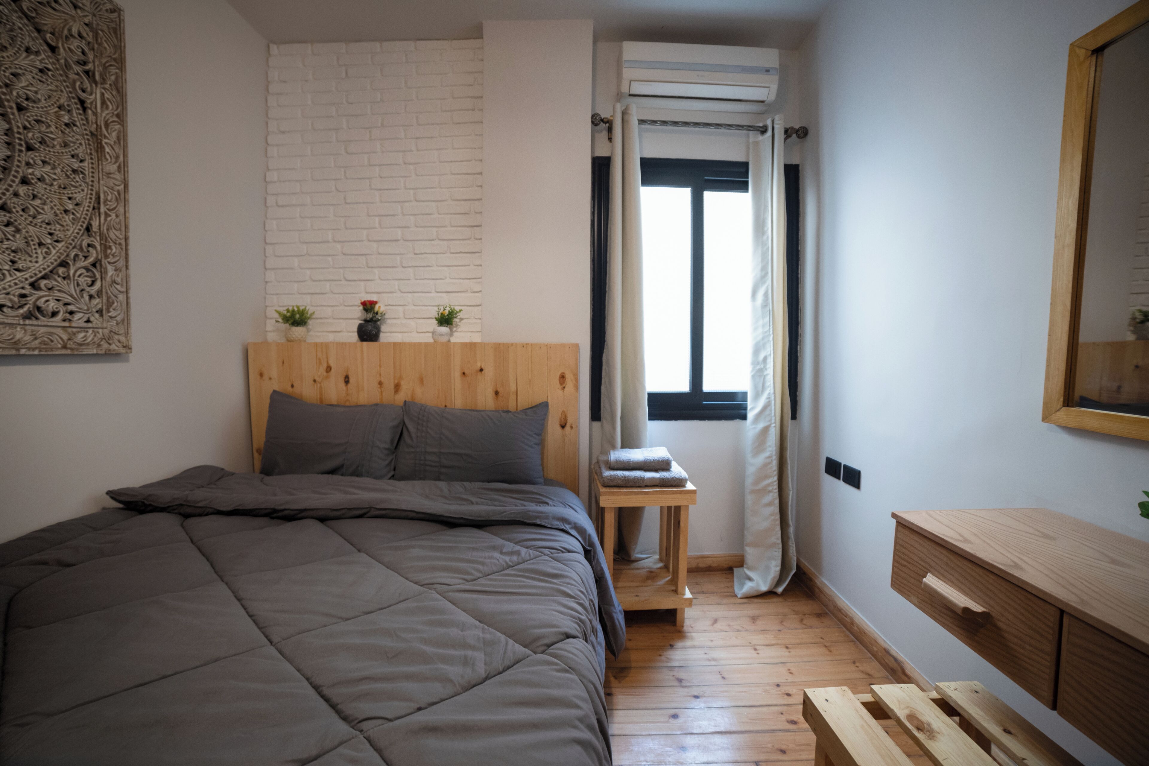 A bedroom with a bed, a wooden headboard, a small table with a towel, a mirror, and a window with curtains.