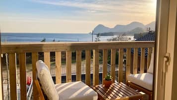 Una terraza de madera con dos sillas acolchadas y una mesita pequeña, con vistas a una playa y montañas al fondo.