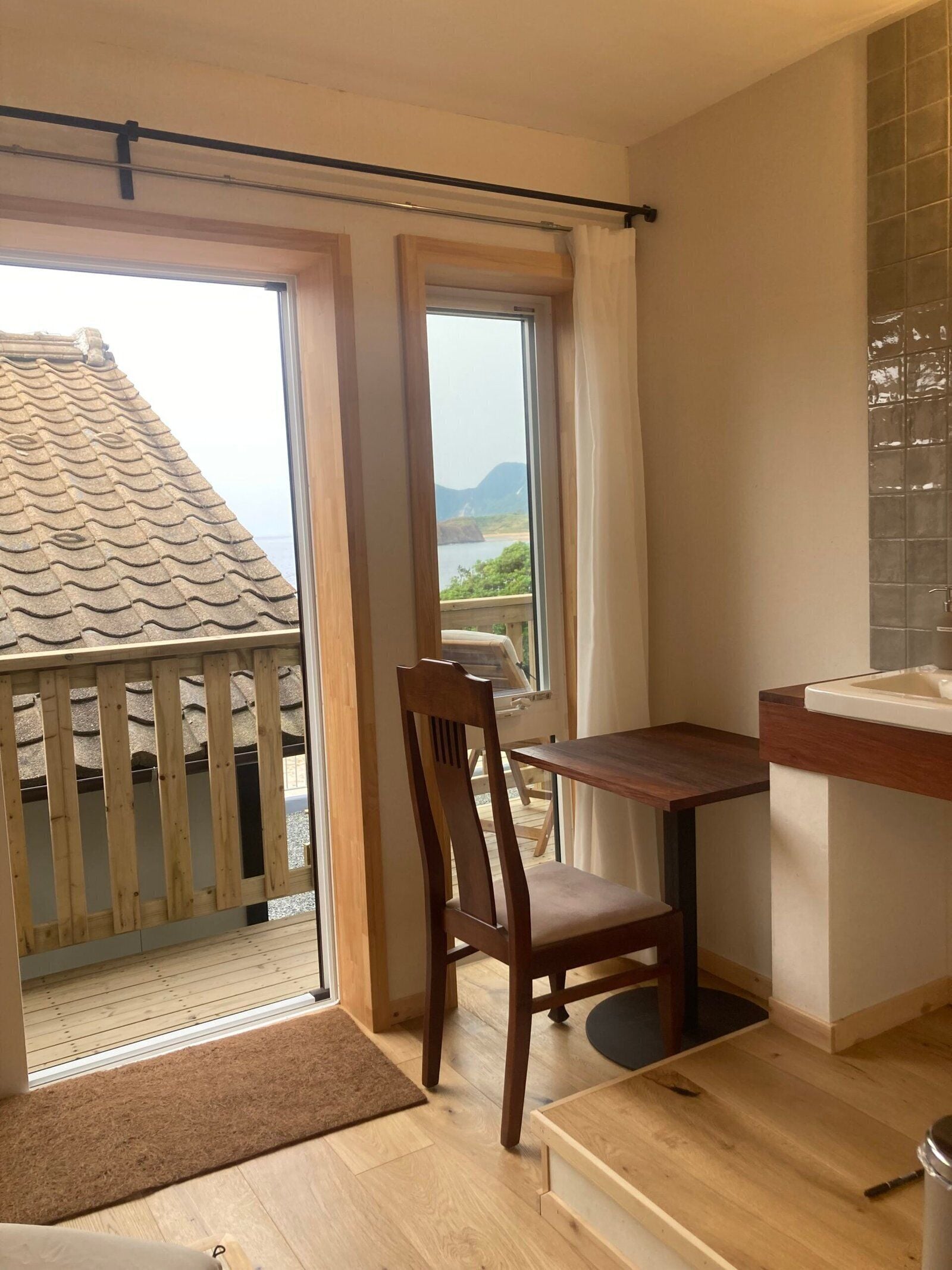 A room with a wooden table, a chair, a window with curtains, a view of the sea, and a tiled wall.