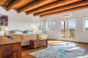 A bedroom with a wooden ceiling, a bed with a headboard, a nightstand, a bedside table, a lamp, a window, and a view of the outdoors. - 1795 Adobe, Dog Friendly · High Road to Taos (Ranchos de Taos)