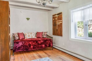A room with a wooden floor, a red sofa with patterned cushions, a wooden door, and a window with a white blind.