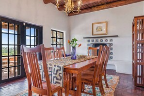 A dining area with a wooden table, chairs, a patterned table runner, a vase with flowers, a fireplace with patterned tiles, and a view through large windows. - 1795 Adobe, Dog Friendly · High Road to Taos (Ranchos de Taos)