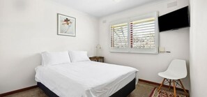 Ein Schlafzimmer mit einem Bett, einem Sessel, einem an der Wand montierten Fernseher und einem Fenster mit Jalousien.