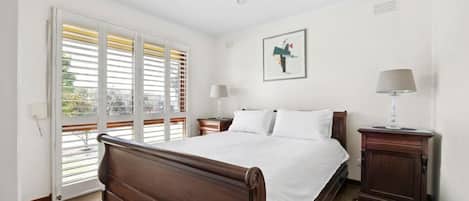 A bedroom with a large bed, two bedside tables, a window with blinds, and a painting on the wall.