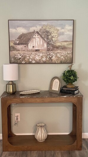 Um console de madeira com um quadro emoldurado acima, uma luminária, um vaso decorativo e uma pequena planta.