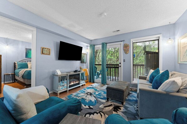 A living room with a fireplace, a flat-screen TV, and a balcony with a view of trees.