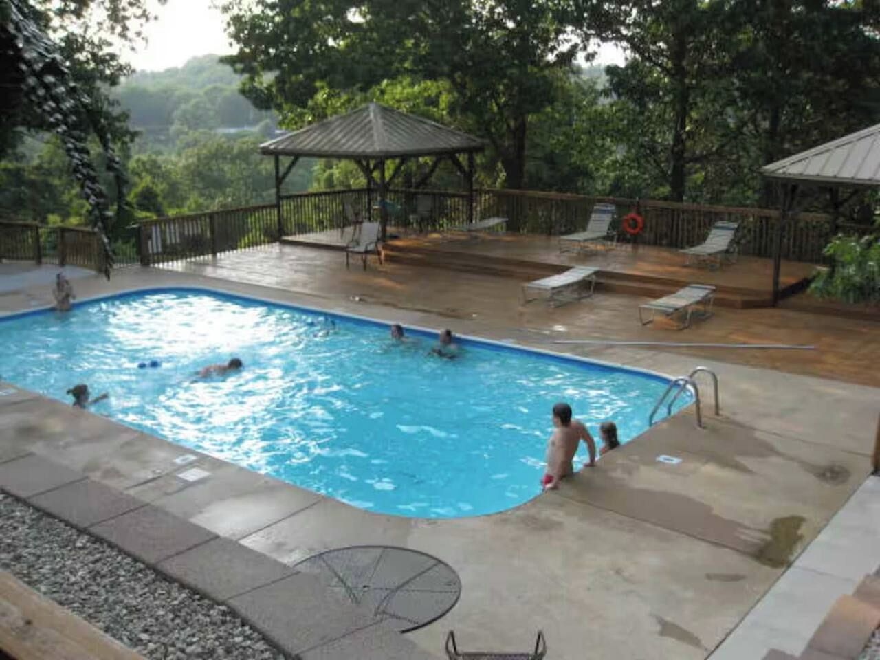 An outdoor swimming pool with people swimming and lounging, surrounded by a deck with chairs and a gazebo.