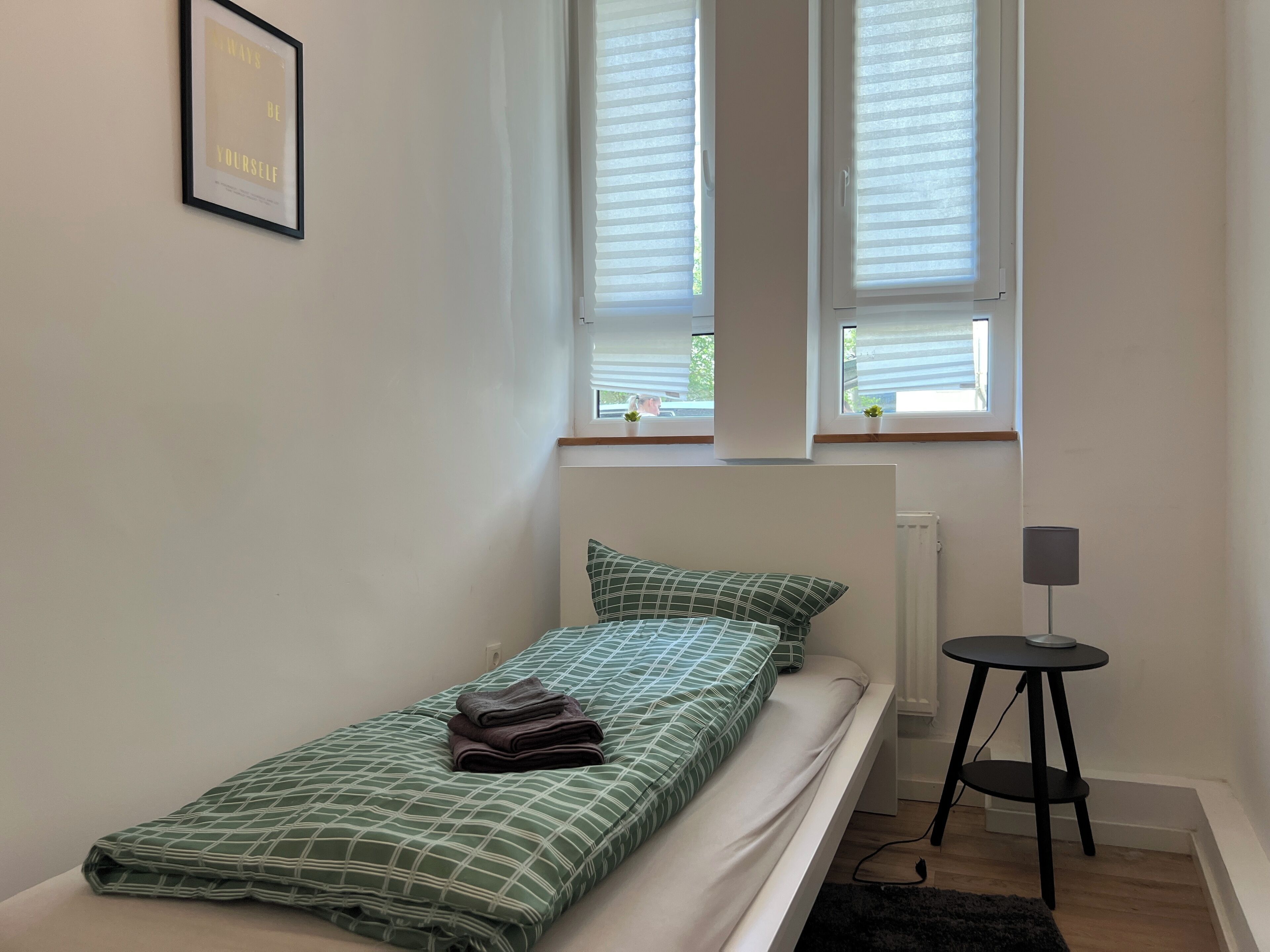 Ein kleines Zimmer mit einem Einzelbett, einem Fenster, einem Heizkörper, einem kleinen Tisch mit Lampe und einem gerahmten Zitat an der Wand.