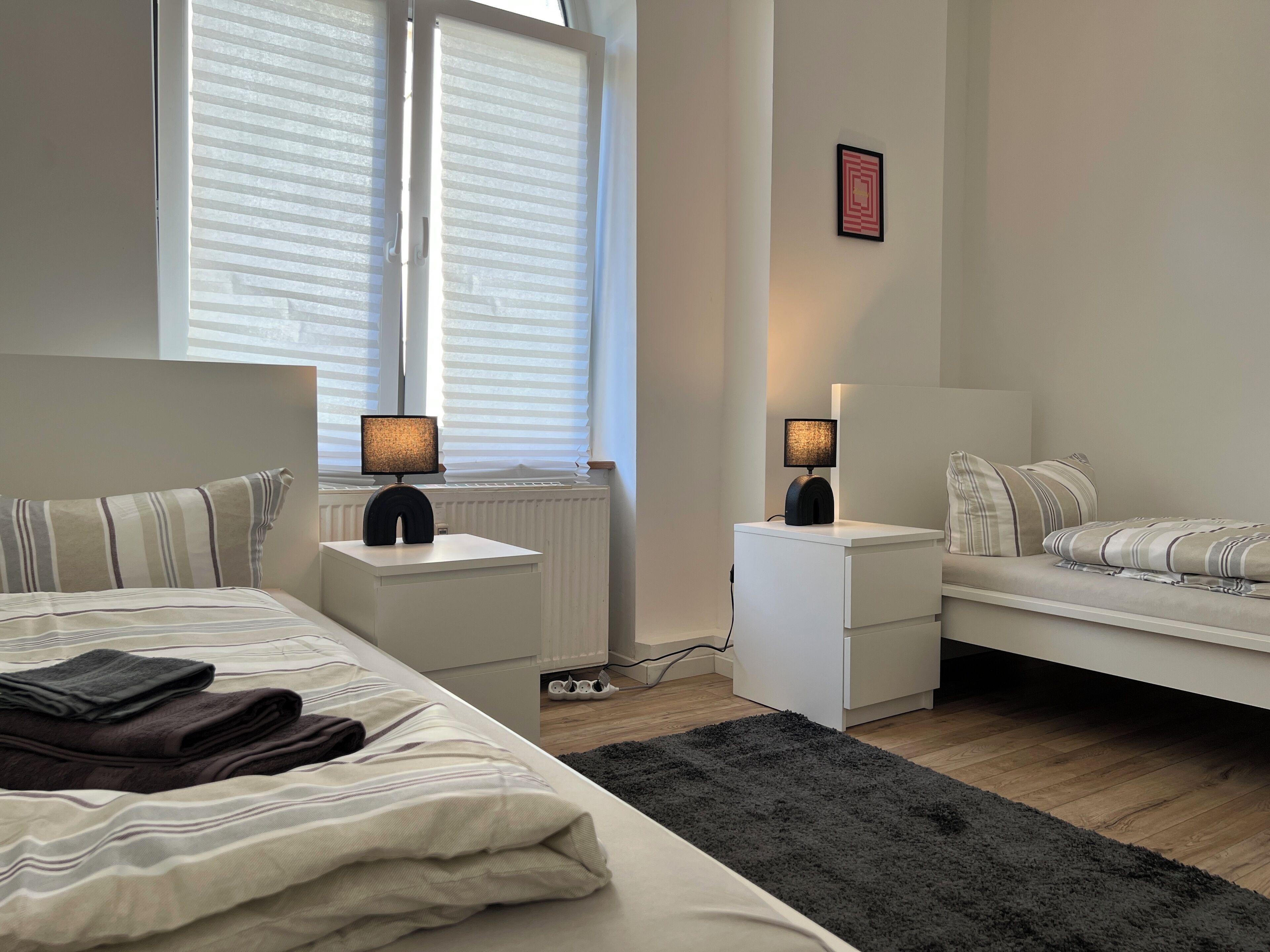 Un dormitorio con cama, mesitas de noche, lámpara de noche, radiador, ventana con persianas, alfombra y un cuadro colgado en la pared.
