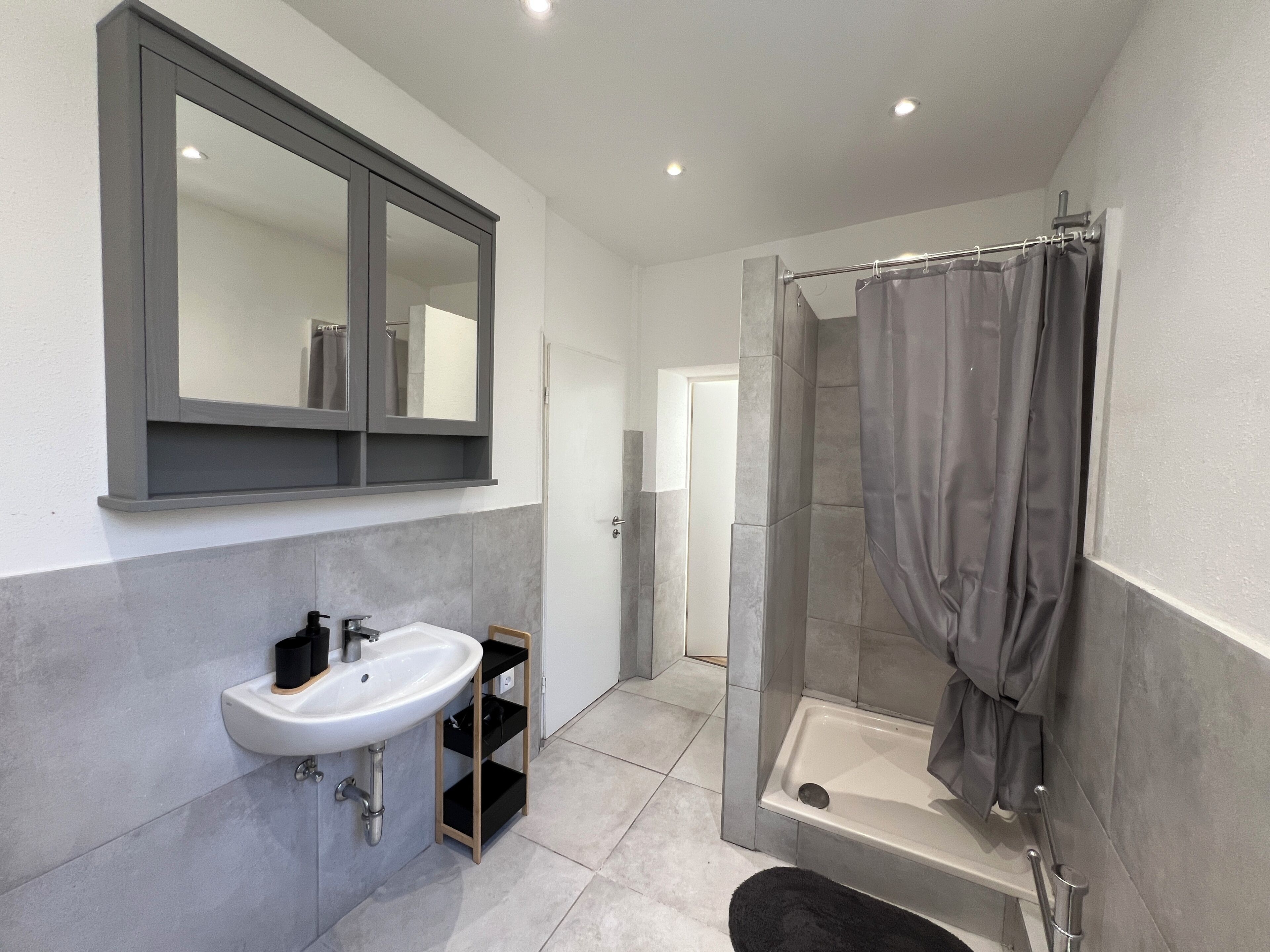 Ein modernes Badezimmer mit Waschbecken, Spiegel, Dusche mit Glastür und grauen Fliesen.