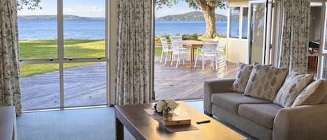 A living room with a large sliding glass door leading to an outdoor deck with a view of the sea and hills.