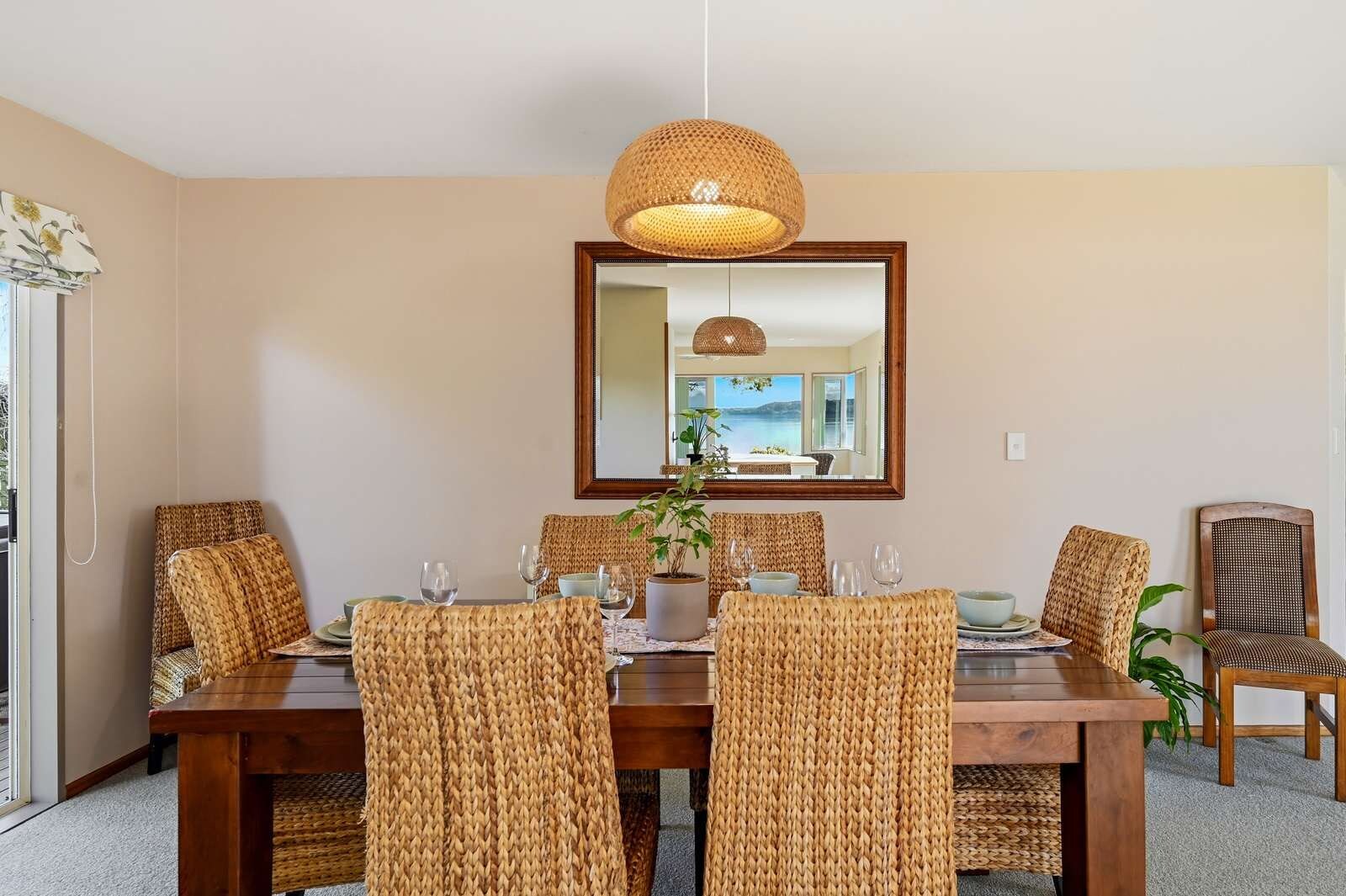 Un espace repas comprenant une table en bois, des chaises en osier, un luminaire suspendu et un grand miroir qui reflète la pièce.