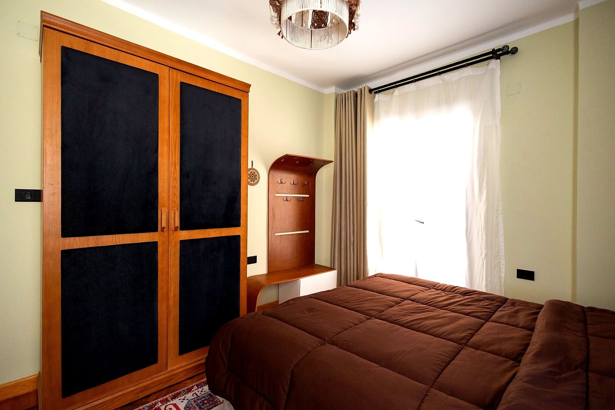 A bedroom with a large wardrobe, a bed with a brown quilt, and a window with curtains.