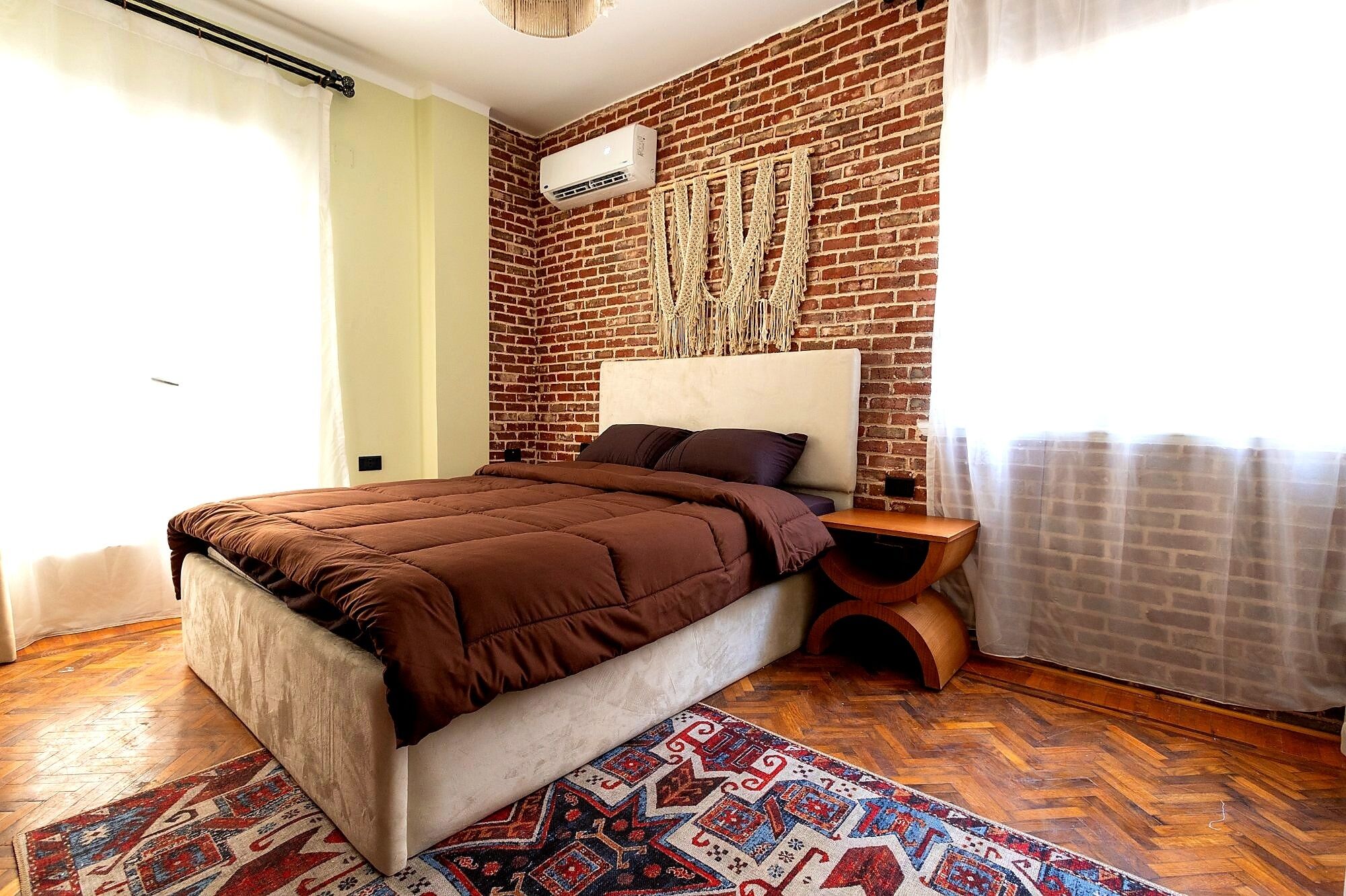 A bedroom with a brick wall, a bed with brown bedding, a headboard, a bedside table with a lamp, and a patterned rug.