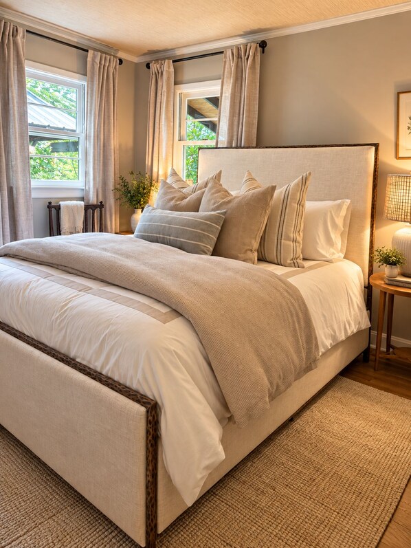A bedroom with a large bed, a bedside table, a lamp, and a view of greenery outside.