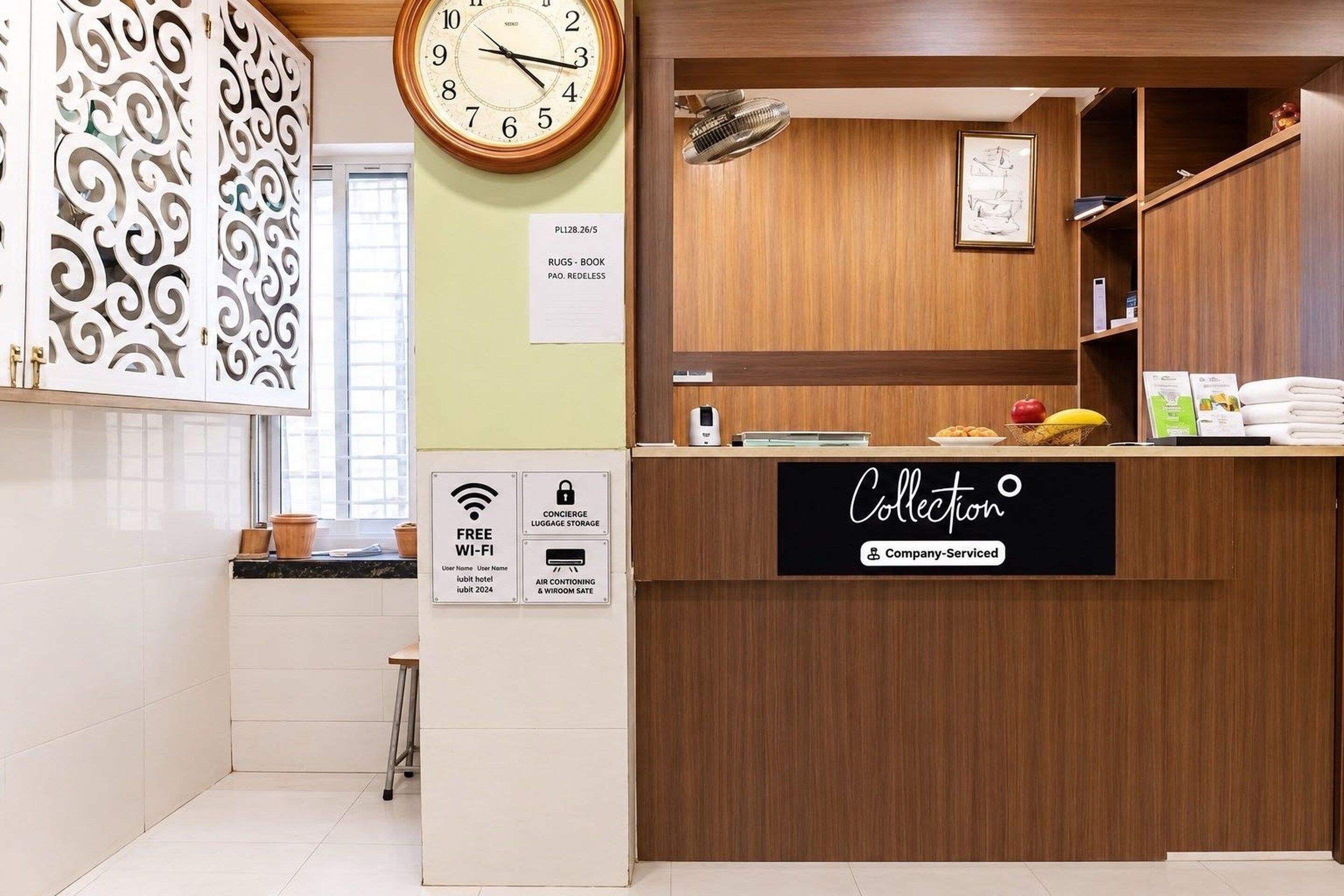 Un espace d’accueil d’hôtel avec un comptoir de réception en bois, une horloge et un panneau indiquant les services proposés, tels que le Wi-Fi gratuit, le service de bagagerie et la climatisation.