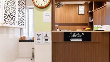 Un espace d’accueil d’hôtel avec un comptoir de réception en bois, une horloge et un panneau indiquant les services proposés, tels que le Wi-Fi gratuit, le service de bagagerie et la climatisation.