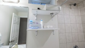 Un baño con lavamanos blanco, espejo, zona de ducha y estante con artículos de aseo.