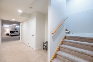 A hallway leading to a bedroom with a bed, a nightstand, and a lamp. There is a white door on the left and a staircase on the right.