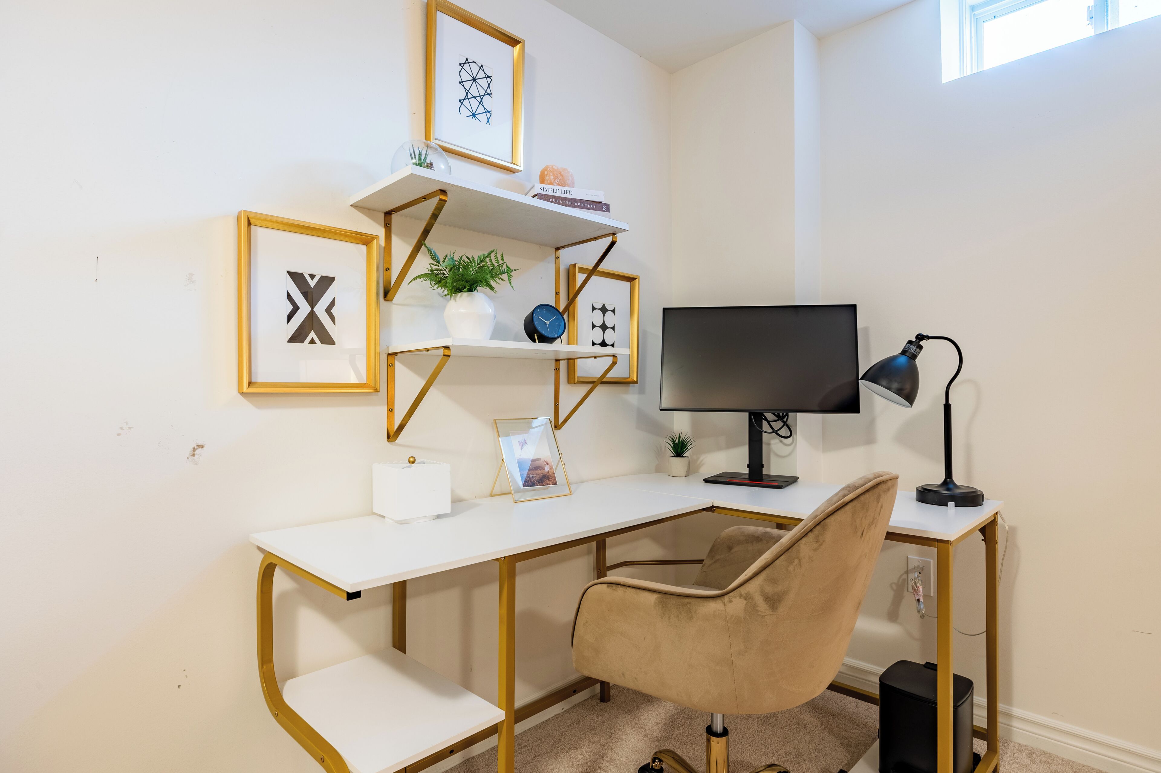 Un escritorio moderno con una computadora, una lámpara y una silla. Hay cuadros enmarcados y una planta pequeña en la pared.