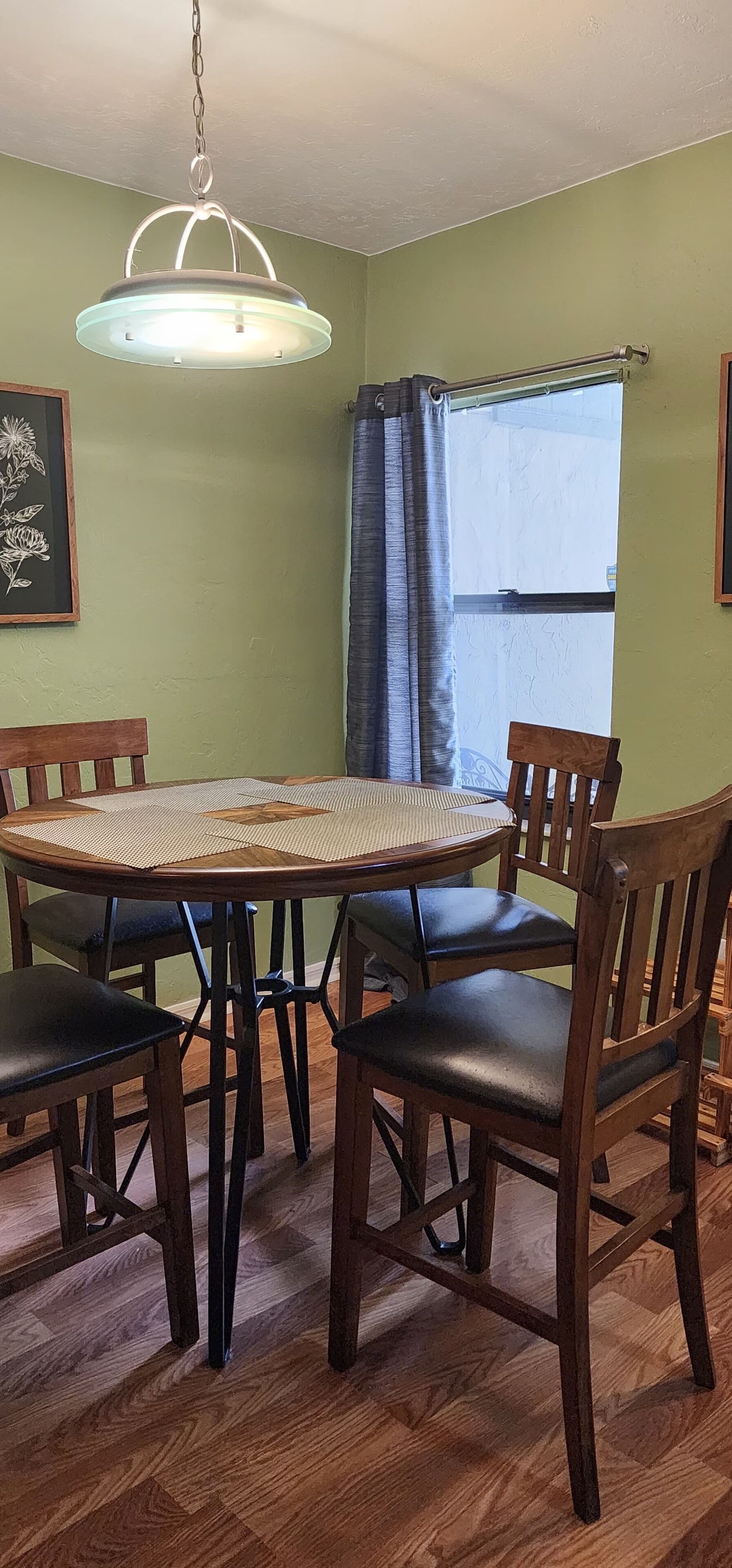 Un coin repas avec une table ronde, quatre chaises, un luminaire suspendu, une fenêtre avec des rideaux et un tableau encadré au mur.