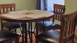 A dining area with a round table, four chairs, a hanging light fixture, a window with curtains, and a framed picture on the wall.