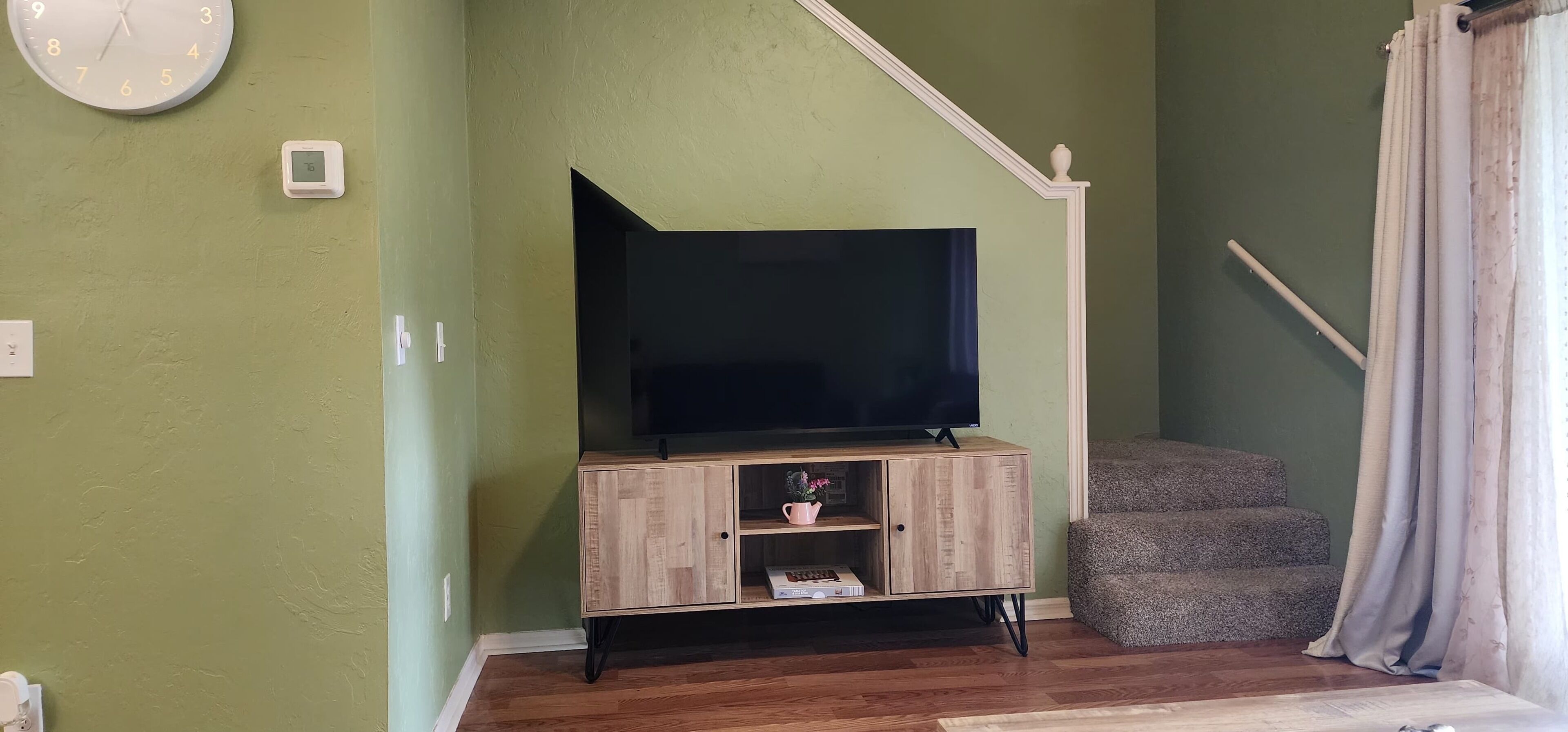A living room with a television on a wooden cabinet, a clock on the wall, and a staircase with a curtain.