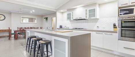 Cozinha moderna com ilha central, banquetas, área de jantar com mesa e cadeiras de madeira, e forno embutido.