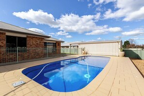 A residential swimming pool with a brick house and a metal fence in the background.