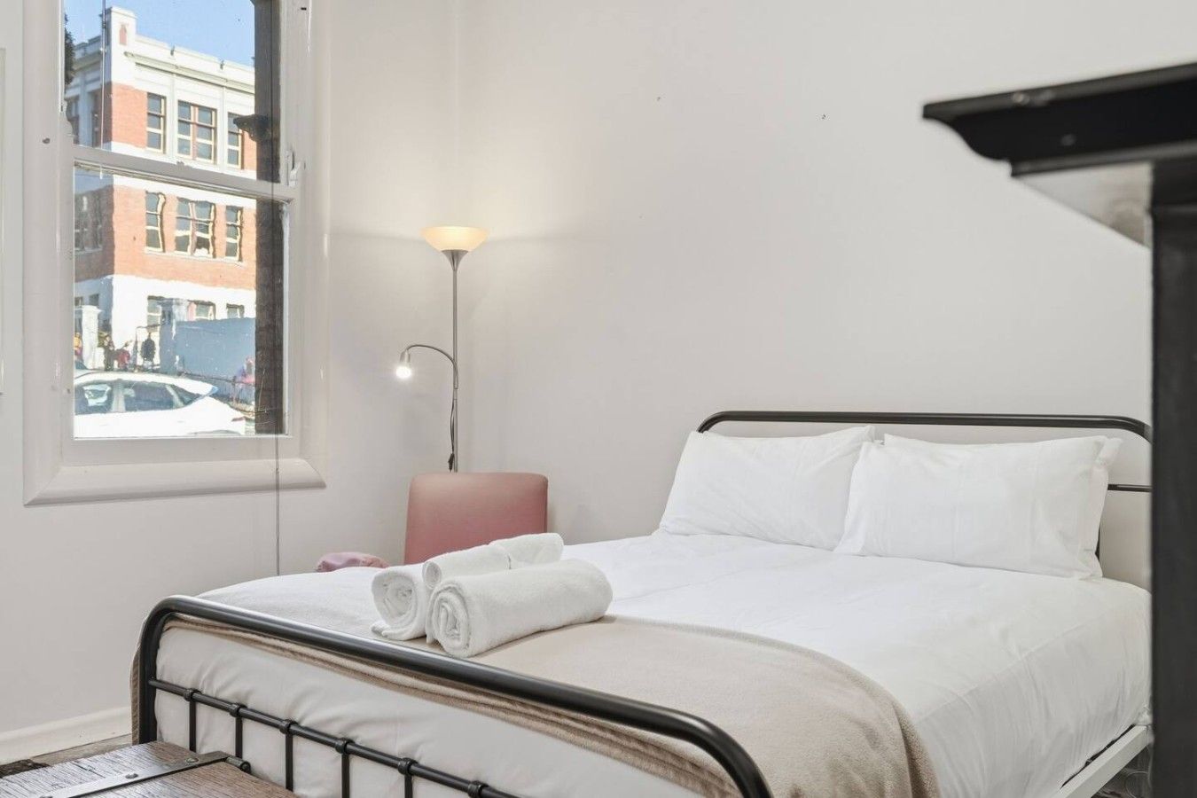 A hotel room with a bed, a bedside lamp, a window with a view of buildings, and a small table.
