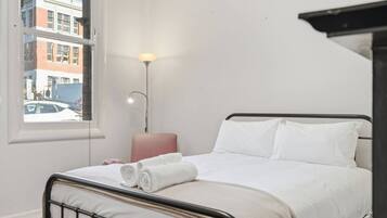A hotel room with a bed, a bedside lamp, a window with a view of buildings, and a small table.