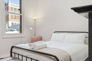 A hotel room with a bed, a bedside lamp, a window with a view of buildings, and a small table. - Edinburgh - Queen Room + Free Parking (Hobart)