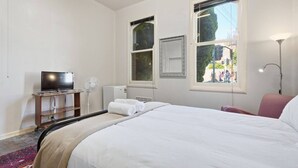 A hotel room with a bed, a television, a fan, a heater, and a window with a view of the street. - Edinburgh - Queen Room + Free Parking (Hobart)