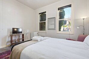 Hotellihuone, jossa on sänky, televisio, tuuletin, lämmitin ja ikkuna, josta on näkymä kadulle.