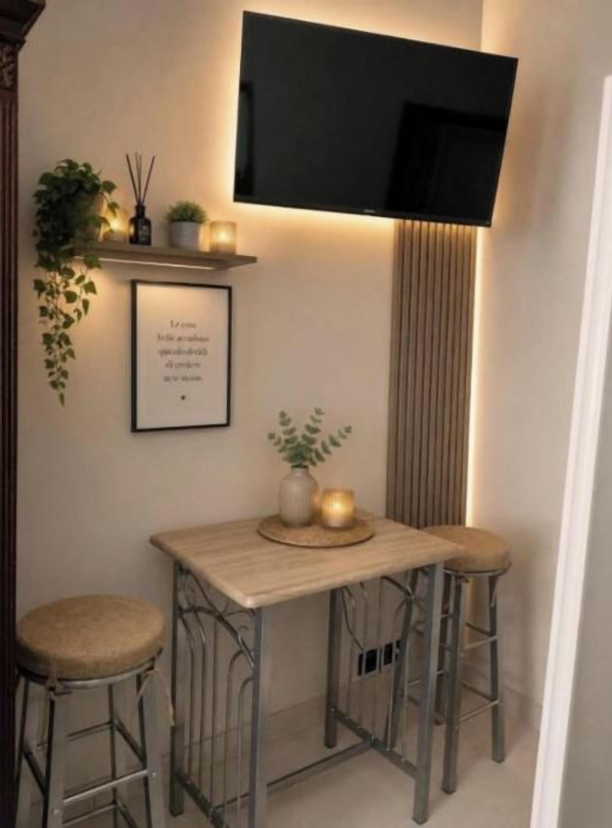 A small, cozy room with a TV mounted on the wall, a wooden table with two stools, and a framed quote on the wall.