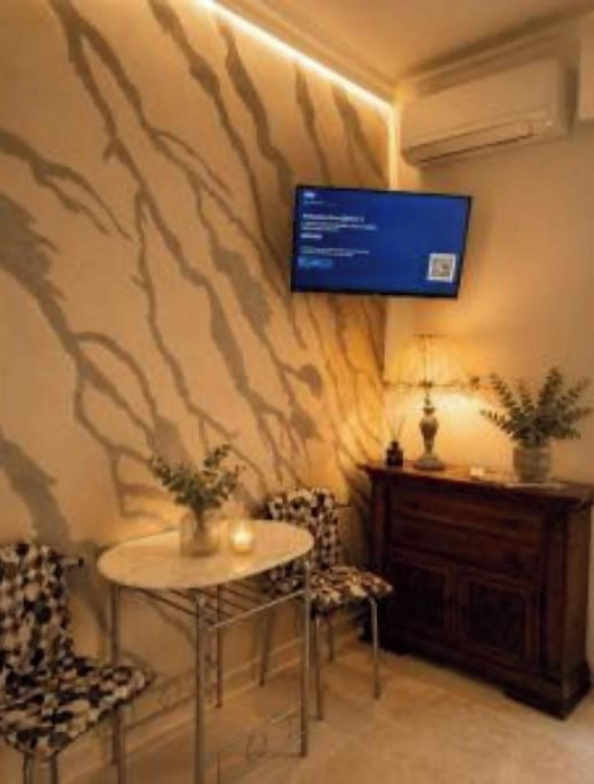 A room with a television mounted on the wall, a round table with chairs, a fireplace, and a patterned wall.